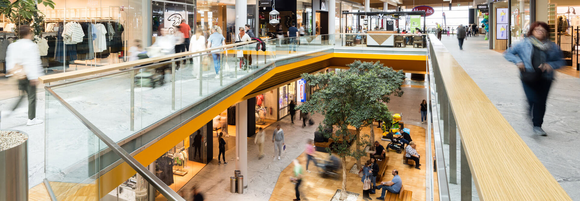 Einkaufszentrum mit vielen Menschen, Geschäften und einem Baum in der Mitte.